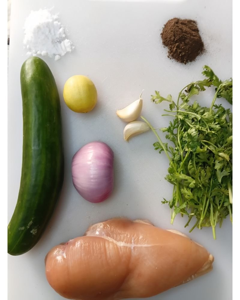 Fresh ingredients for a healthy 5 ingredient chicken salad recipe including cooked chicken breast, cucumber, onion, cilantro, lemon, and simple seasonings arranged on a wooden countertop.